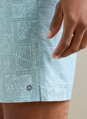 A close-up of a hand rests beside Peter Millar’s Seaside Shade Swim Trunk, featuring a light blue leaf pattern, small circular logo, and an elastic drawstring waistband for adjustable comfort.