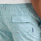 Close-up of a person wearing Peter Millar Seaside Shade Swim Trunk featuring an elastic waistband, flap pocket, metal grommets, and quick-drying light blue patterned fabric.