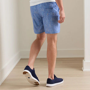 A man stands indoors on a light wood floor, facing away, wearing Peter Millar Tequila Seas Swim Trunk with an elastic drawstring waist, a white T-shirt, and navy slip-on shoes.