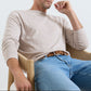 A man sits in a wooden chair wearing the Peter Millar Lake Long Sleeve Sweater in light beige and blue jeans, with one arm resting on the chair and his other hand near his face.