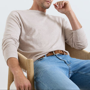 A man sits in a wooden chair wearing the Peter Millar Lake Long Sleeve Sweater in light beige and blue jeans, with one arm resting on the chair and his other hand near his face.