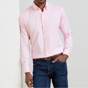 A person in a light pink Peter Millar Campbell Sport Shirt, blue jeans, and a brown belt stands with one hand in their pocket against a light background.