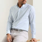 A man wearing the Peter Millar Rommel Performance Twill Sport Shirt in light blue check with light beige pants sits on a wooden stool against a plain background.