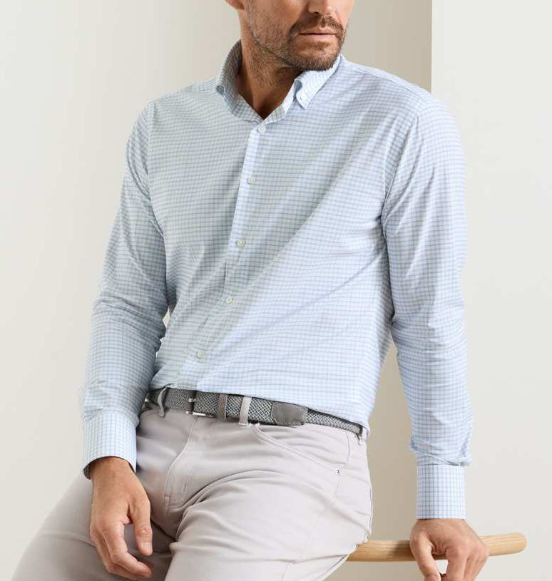 A man wearing the Peter Millar Rommel Performance Twill Sport Shirt in light blue check with light beige pants sits on a wooden stool against a plain background.