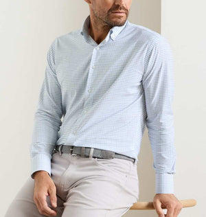 A man wearing the Peter Millar Rommel Performance Twill Sport Shirt in light blue check with light beige pants sits on a wooden stool against a plain background.