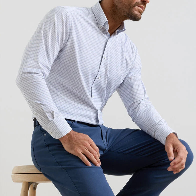 A man sits on a stool in the Peter Millar Rommel Performance Twill Sport Shirt—a light checked shirt—paired with blue pants, in front of a plain background.