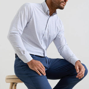 A man sits on a stool in the Peter Millar Rommel Performance Twill Sport Shirt—a light checked shirt—paired with blue pants, in front of a plain background.