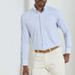 A man wearing the Peter Millar Casey Performance Twill Sport Shirt in light blue check, paired with beige pants and a tan belt, stands against a plain light-colored background.