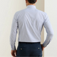 A man is seen from behind wearing the Peter Millar Phillip Performance Twill Sport Shirt in light blue checks and navy dress pants, standing before a light-colored wall.