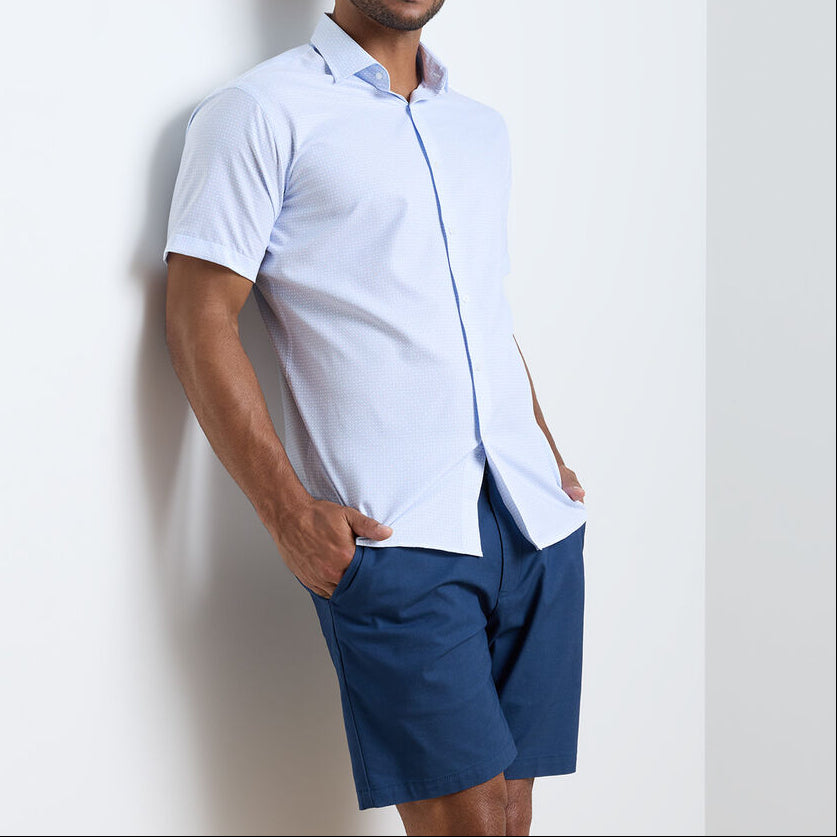 A man stands against a white wall wearing a light blue Peter Millar Baldwin Performance Sport Shirt and dark blue shorts, hands in his pockets.