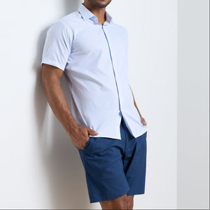 A man stands against a white wall wearing a light blue Peter Millar Baldwin Performance Sport Shirt and dark blue shorts, hands in his pockets.