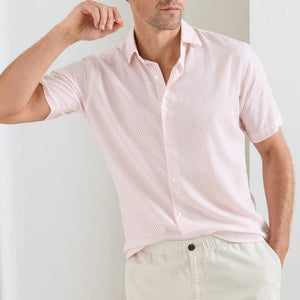 A man wearing a light pink Peter Millar Tamarama Cotton Lyocell Sport Shirt and beige pants stands with one arm bent near his head.