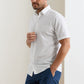 A man wears the Peter Millar Seaward Cotton Lyocell Sport Shirt, short-sleeve and white, paired with blue pants. He stands with one hand in his pocket against a plain background.
