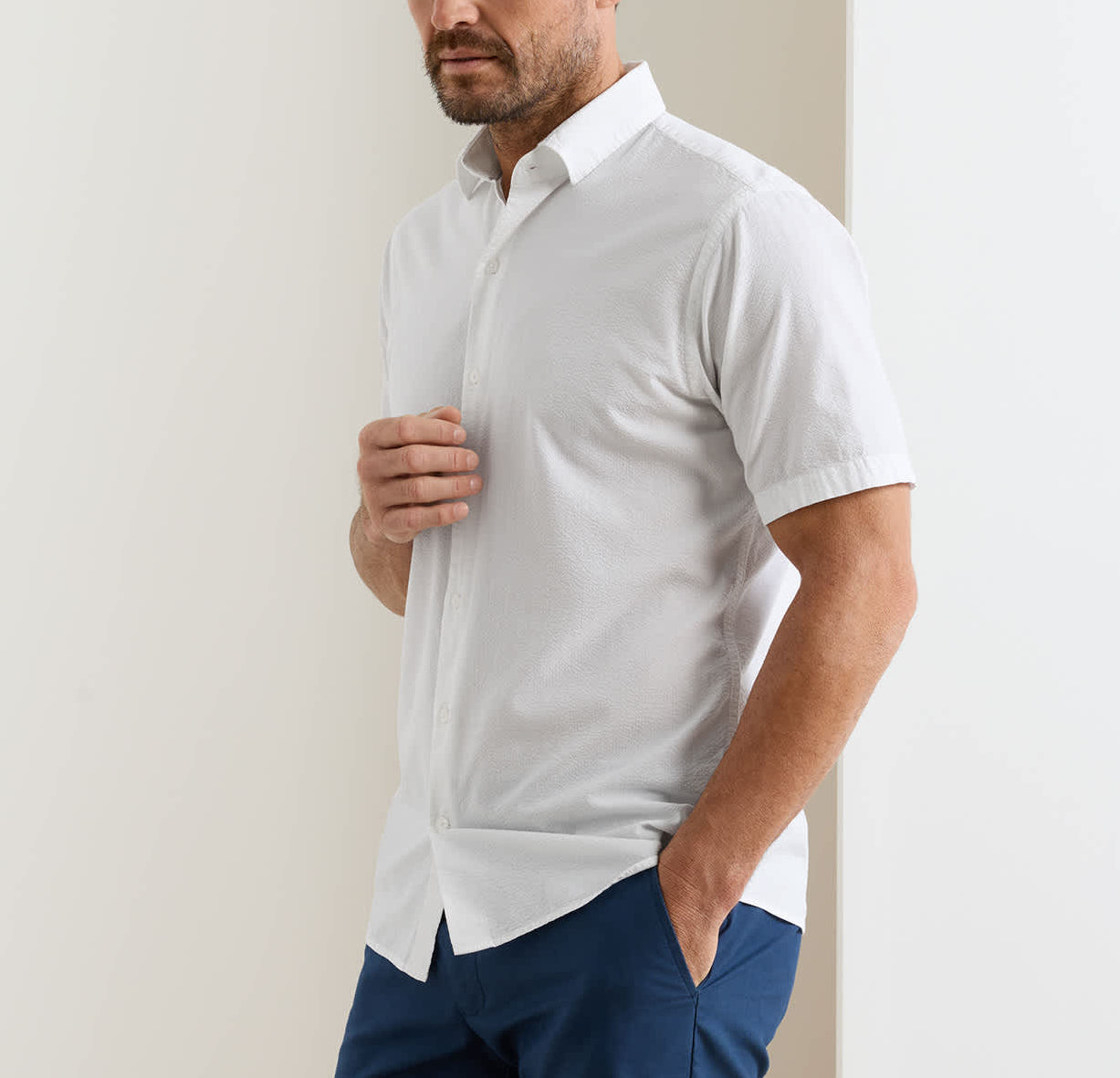 A man wears the Peter Millar Seaward Cotton Lyocell Sport Shirt, short-sleeve and white, paired with blue pants. He stands with one hand in his pocket against a plain background.