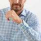 A man in a Peter Millar Waterrock Cotton Lyocell Sport Shirt in light blue and white plaid sits with his hand near his face, revealing part of his chest and lower face.