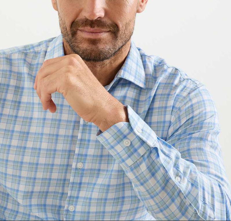 A man in a Peter Millar Waterrock Cotton Lyocell Sport Shirt in light blue and white plaid sits with his hand near his face, revealing part of his chest and lower face.
