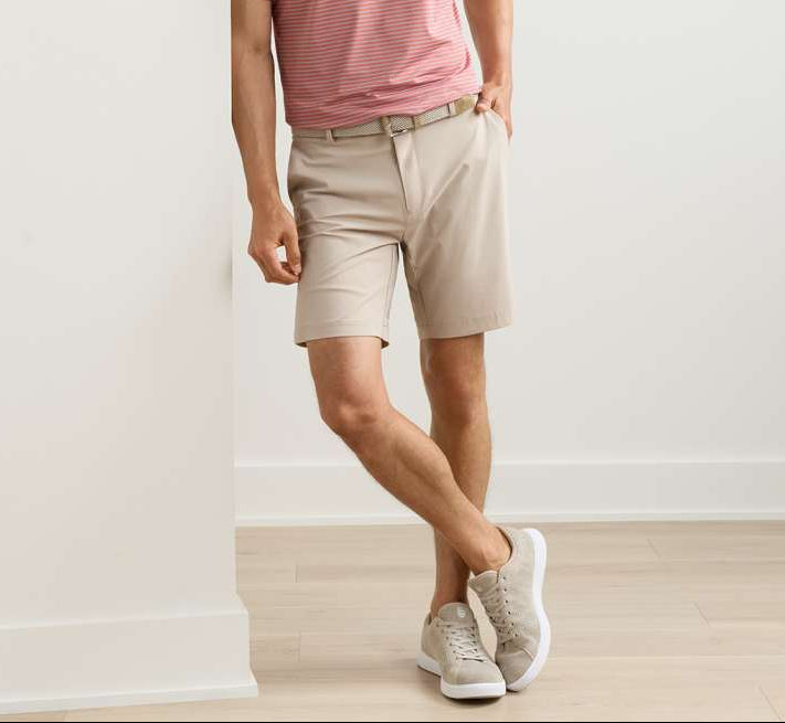 A person wears a red striped shirt, Peter Millar Bingham Performance Short, and white sneakers while standing indoors against a white wall with one leg crossed.