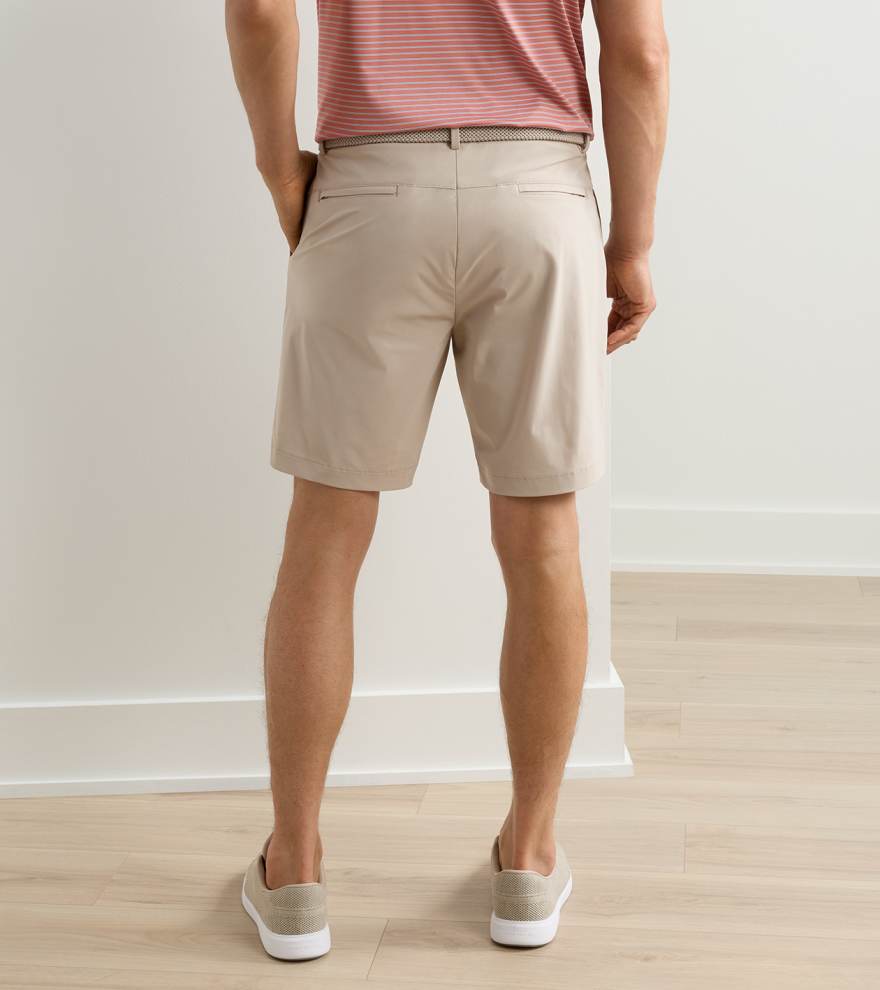 A person wearing Peter Millar Bingham Performance Shorts, a brown belt, red striped shirt, and light slip-on shoes stands facing away in a room with light-colored floors and walls.