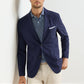 A man wearing the Peter Millar Bingham Performance Blazer over a blue and white striped polo shirt with beige pants, standing against a white wall.