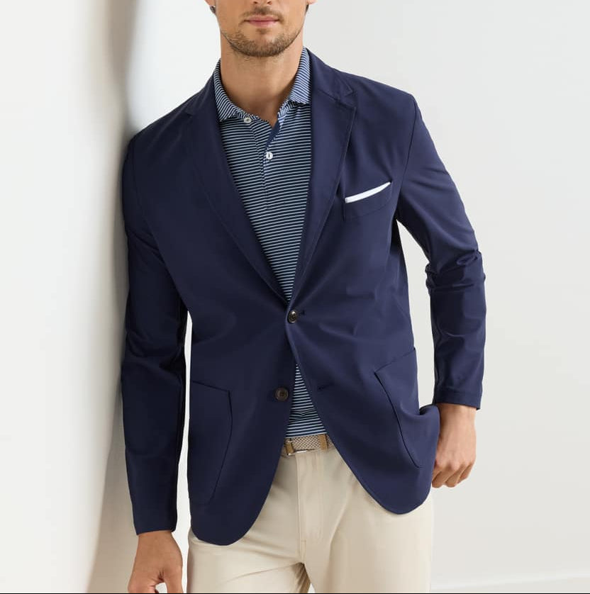 A man wearing the Peter Millar Bingham Performance Blazer over a blue and white striped polo shirt with beige pants, standing against a white wall.