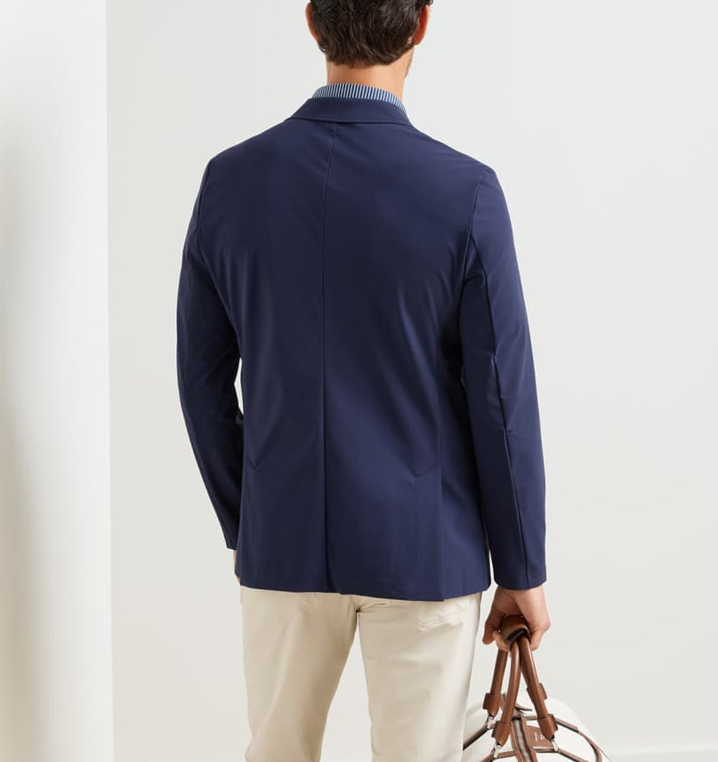 A man is seen from behind wearing the Peter Millar Bingham Performance Blazer in navy with beige pants, holding a brown-handled bag in his left hand against a plain background.