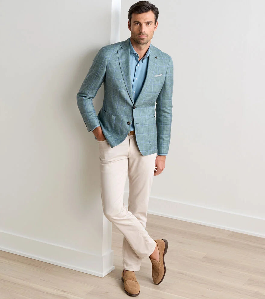 A man stands indoors against a white wall, wearing the Peter Millar Salisbury Windowpane Soft Jacket—a crafted essential—paired with a blue shirt, beige pants, and brown loafers, with one leg casually crossed in front of the other.