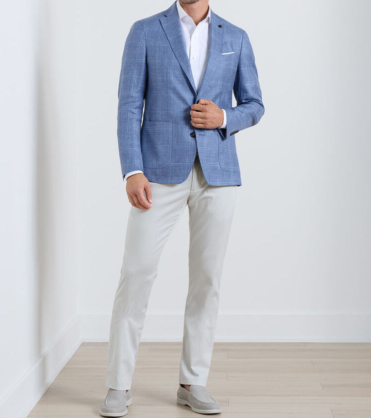 A man wearing the Peter Millar Reynolds Plaid Soft Jacket stands against a white wall and wooden floor, showcasing this light blue lightweight wool blazer as a versatile, casually refined wardrobe essential.