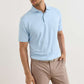 A man stands with one hand in his pocket against a plain background, wearing Peter Millar's Keynote Performance Jersey Polo in light blue and beige pants with a woven belt.