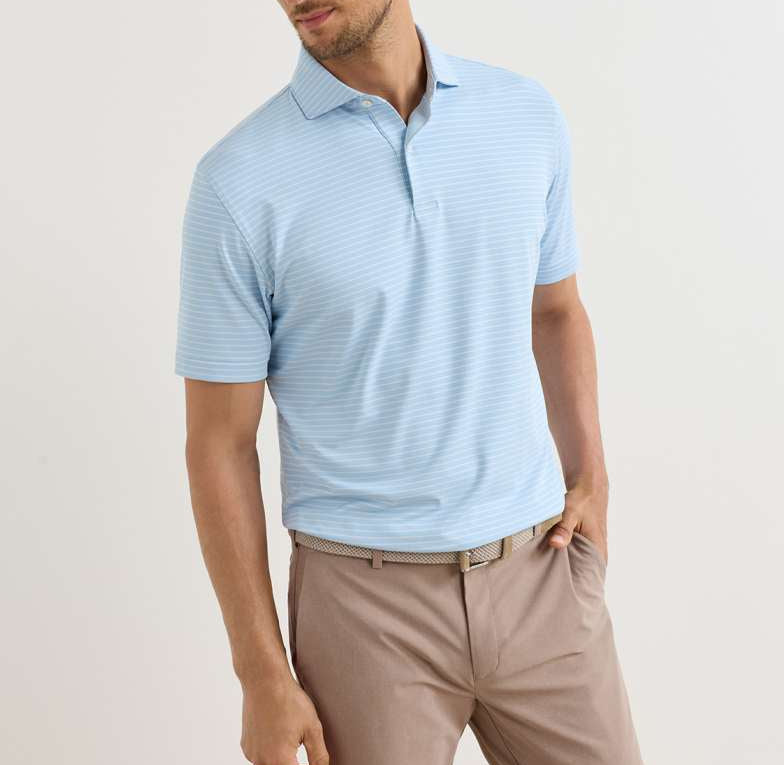 A man stands with one hand in his pocket against a plain background, wearing Peter Millar's Keynote Performance Jersey Polo in light blue and beige pants with a woven belt.