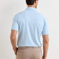 A man stands facing away, wearing a light blue Peter Millar Keynote Performance Jersey Polo with four-way stretch and beige pants with a woven belt.
