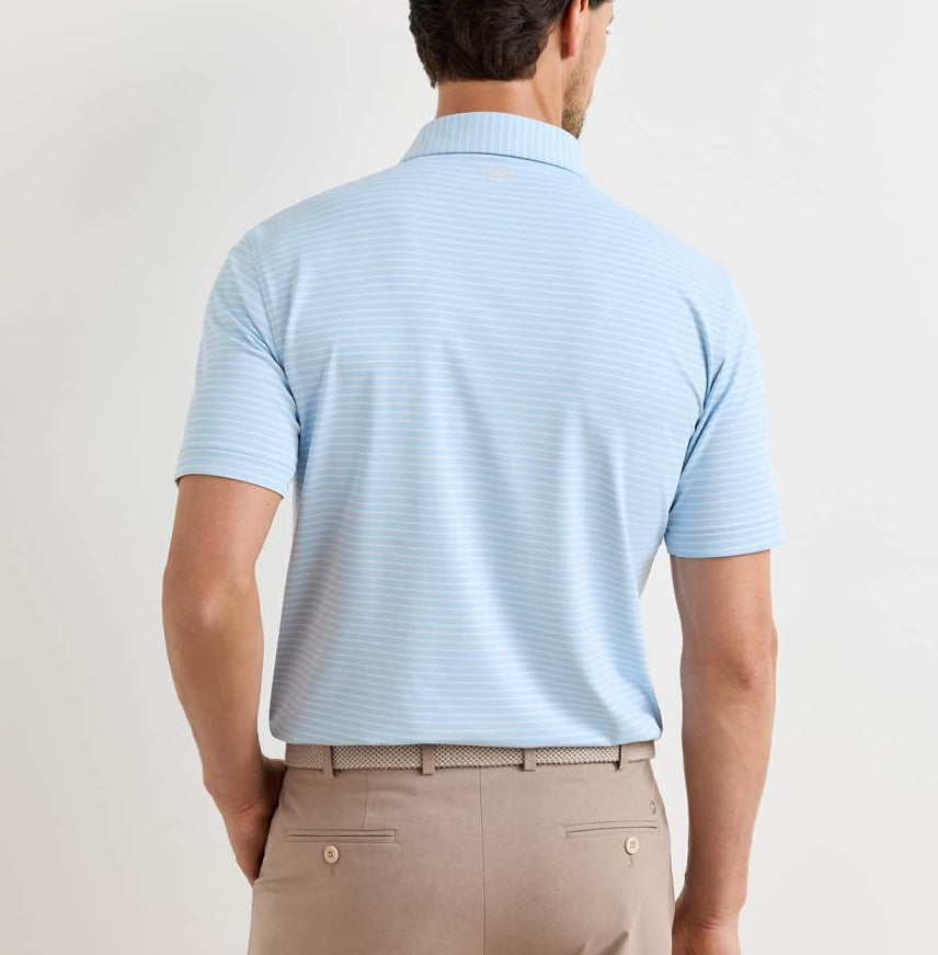 A man stands facing away, wearing a light blue Peter Millar Keynote Performance Jersey Polo with four-way stretch and beige pants with a woven belt.
