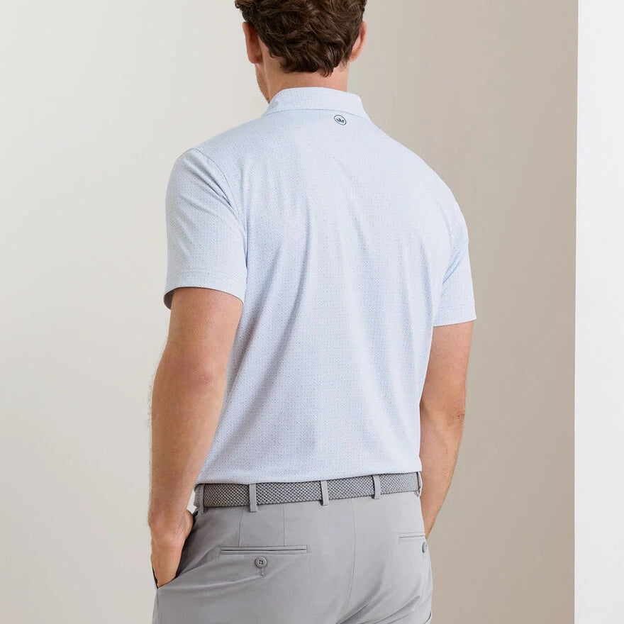 A man stands with his back to the camera in a minimal indoor setting, wearing the Peter Millar Alder Performance Jersey Polo in light blue and light gray pants.