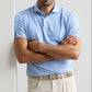 A man stands with arms crossed wearing the Peter Millar Granada Performance Jersey Polo in light blue, featuring four-way stretch and UPF 50+ sun protection, paired with beige pants and a light-colored woven belt against a plain background.