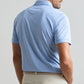 A man shown from the back against a plain background wears the Peter Millar Granada Performance Jersey Polo in light blue with short sleeves and four-way stretch, paired with beige pants.