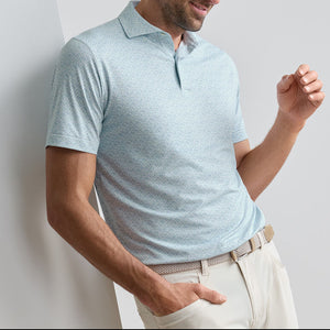 A man stands against a white wall wearing the Peter Millar Wood Fired Performance Jersey Polo in light blue with UPF 50+ sun protection and beige pants, one arm relaxed.