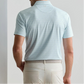 A man is seen from behind wearing a Peter Millar Wood Fired Performance Jersey Polo in light blue, featuring four-way stretch, tucked into beige pants with a textured belt.