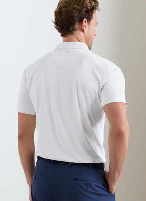 A man stands facing away, wearing the Peter Millar Fermata Performance Jersey Polo with short sleeves and four-way stretch, paired with navy blue pants.