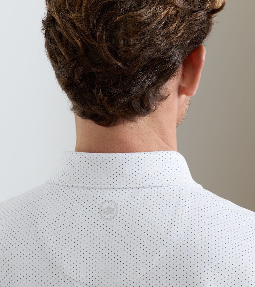 A man with brown hair seen from behind wears the Peter Millar Fermata Performance Jersey Polo, a white collared shirt with small black dots, four-way stretch, and a circular logo printed below the collar on the back.