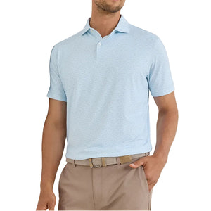 A man wears the Peter Millar Soprano Waves Performance Jersey Polo in light blue (UPF 50+), tucked into beige pants with a woven belt, standing with one hand in his pocket.