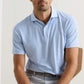 A man wears a Peter Millar Mayfield Performance Jersey Polo in light blue with UPF 50+ and light gray pants, sitting on a wooden chair against a plain background.