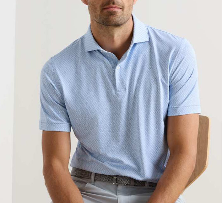 A man wears a Peter Millar Mayfield Performance Jersey Polo in light blue with UPF 50+ and light gray pants, sitting on a wooden chair against a plain background.