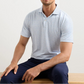 A man wearing a light blue Peter Millar Melody Sea Performance Jersey Polo (UPF 50+, short-sleeve) and navy pants sits on a wooden bench against a plain background.