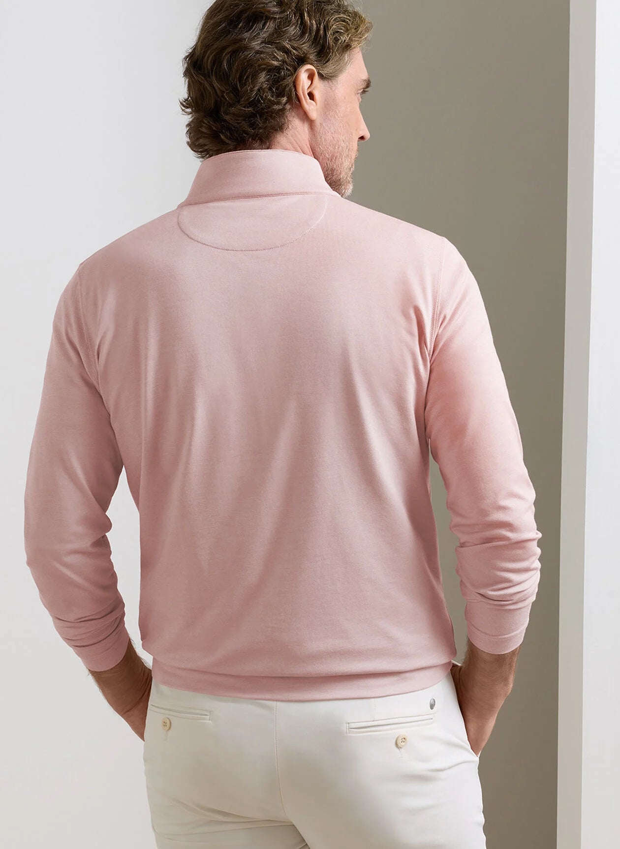 A man, seen from behind in a minimal indoor setting, wears the Peter Millar Albatross Cotton-Blend Piqué Quarter-Zip in light pink paired with white pants.