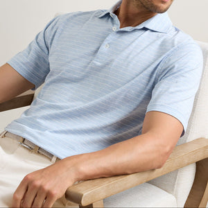 A man sits in a wooden chair wearing a light blue, short-sleeved Peter Millar Excursionist Flex Striped Polo with thin white stripes, made from cotton jersey knit, and beige pants.