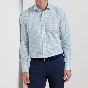 A man wearing the Peter Millar Tropea Italian Poplin Sport Shirt in light blue plaid and navy blue pants stands against a white wall.