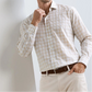 A man wearing the Peter Millar Venzone Italian Poplin Sport Shirt in light beige plaid, paired with light khaki pants and a brown belt, stands with one hand in his pocket and the other touching his chin.