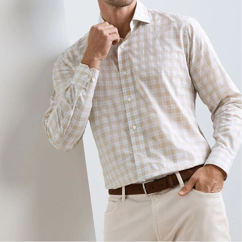 A man wearing the Peter Millar Venzone Italian Poplin Sport Shirt in light beige plaid, paired with light khaki pants and a brown belt, stands with one hand in his pocket and the other touching his chin.