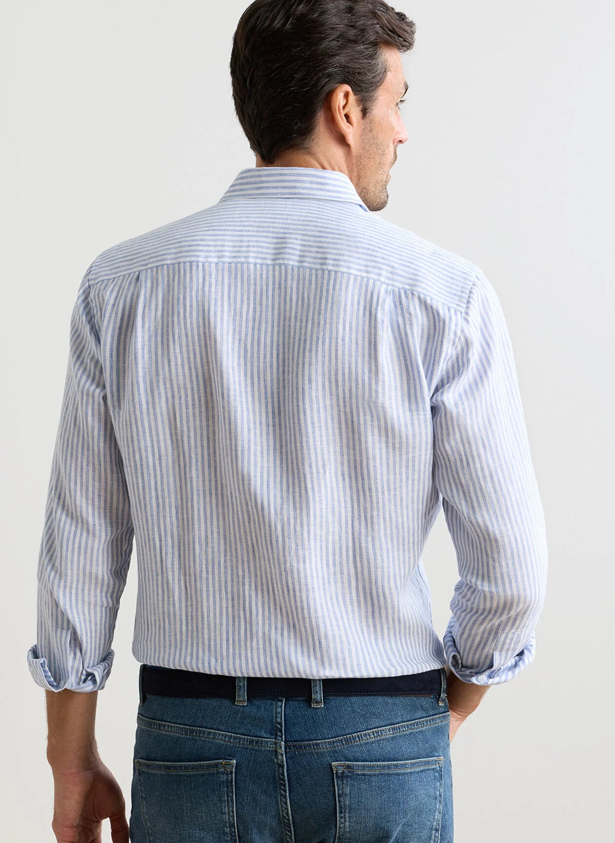 A man is shown from the back wearing the Peter Millar Astino Lino Stripe Sport Shirt in light blue and white Italian linen with mother-of-pearl buttons, paired with blue jeans, against a plain background.