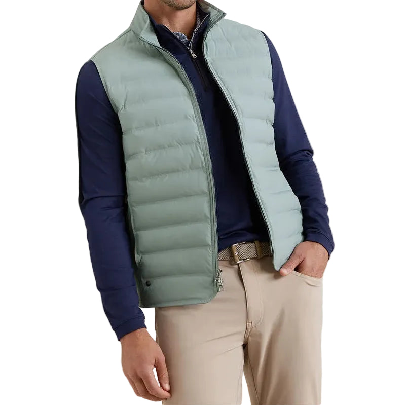 A man wears the Peter Millar Winsome Hybrid Vest in light green, featuring channel insulation and a two-way zip, layered over a navy long-sleeve shirt and beige pants, with one hand in his pocket.