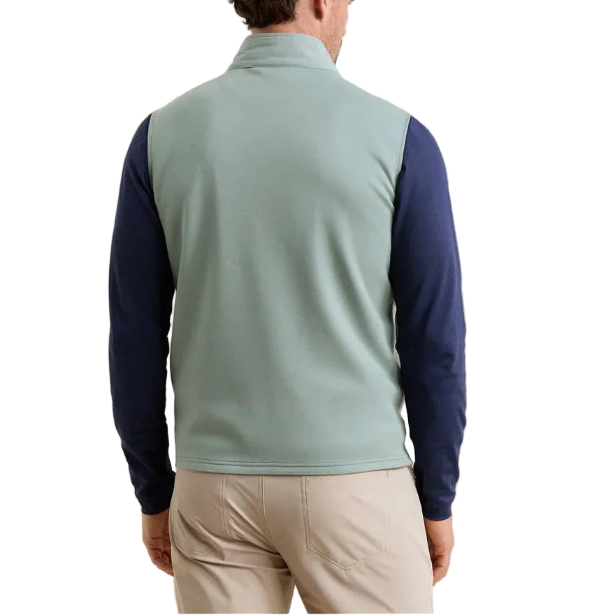 A man stands facing away, wearing the Peter Millar Winsome Hybrid Vest in light green over a navy long-sleeve shirt and beige pants against a white background.